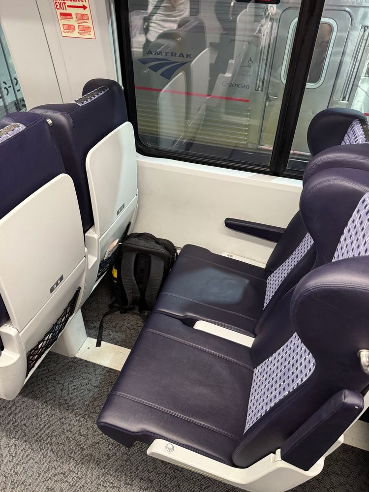 Interior view of a train seating area featuring two adjacent seats with a dark leather-like covering. A black backpack is placed on the floor nearby. The window shows an "Amtrak" logo and a hint of the platform outside.