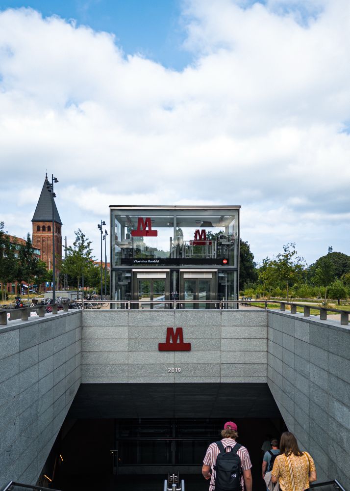 A modern subway entrance with glass construction and a large red "M" symbol is shown. Two people are walking down the stairs, and a brick tower can be seen in the background. The setting has greenery and a cloudy blue sky.