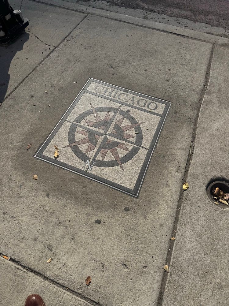 A mosaic compass design embedded in a sidewalk, featuring the word "Chicago" at the top and directional markers. Some leaves are scattered around the area.