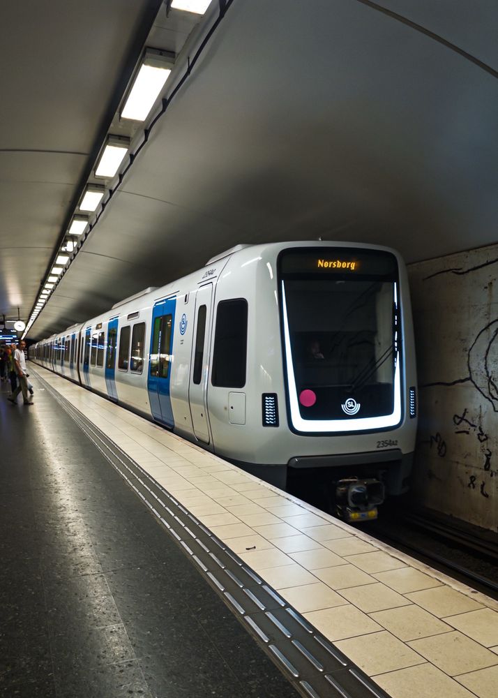 A modern subway train is stationed at a platform inside a well-lit underground station. The train has a white and blue exterior, with "Norsborg" displayed on the front. There are people waiting near the platform, and the surrounding walls feature drawings carved into them