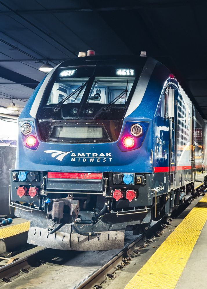 A blue Amtrak train labeled "Midwest" is parked on tracks within a station. The front view shows the train's headlights, logo, and various details of the engineering cabin. The platform features yellow safety markings.