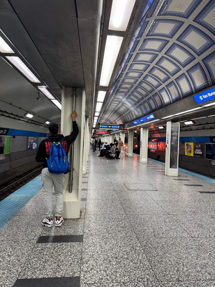 Jackson Blue Line CTA station platform featuring a man standing near a support pillar, holding onto it. He is wearing a black jacket with colorful text, gray pants, and red sneakers, with a blue backpack. In the background, several people are seated waiting for the train