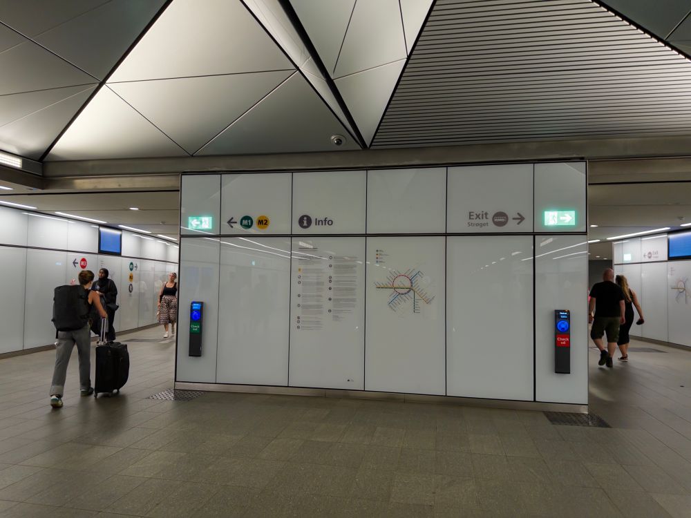 A modern subway station interior with directional signage for train lines (M1, M2) and an information display. An exit sign points to "Strøget." Passengers with luggage and casual attire are visible, along with a map of the transit system