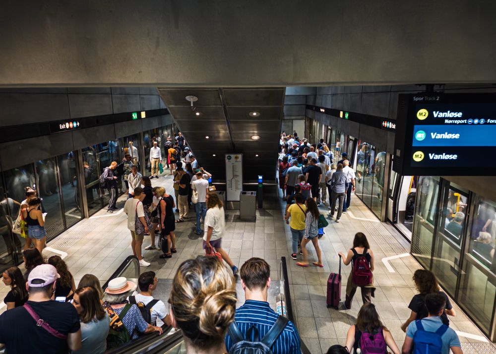 A crowded subway station showing people waiting and boarding trains. Signs indicate the direction to "Vanløse," and the platform has a modern design with escalators and clear signage.