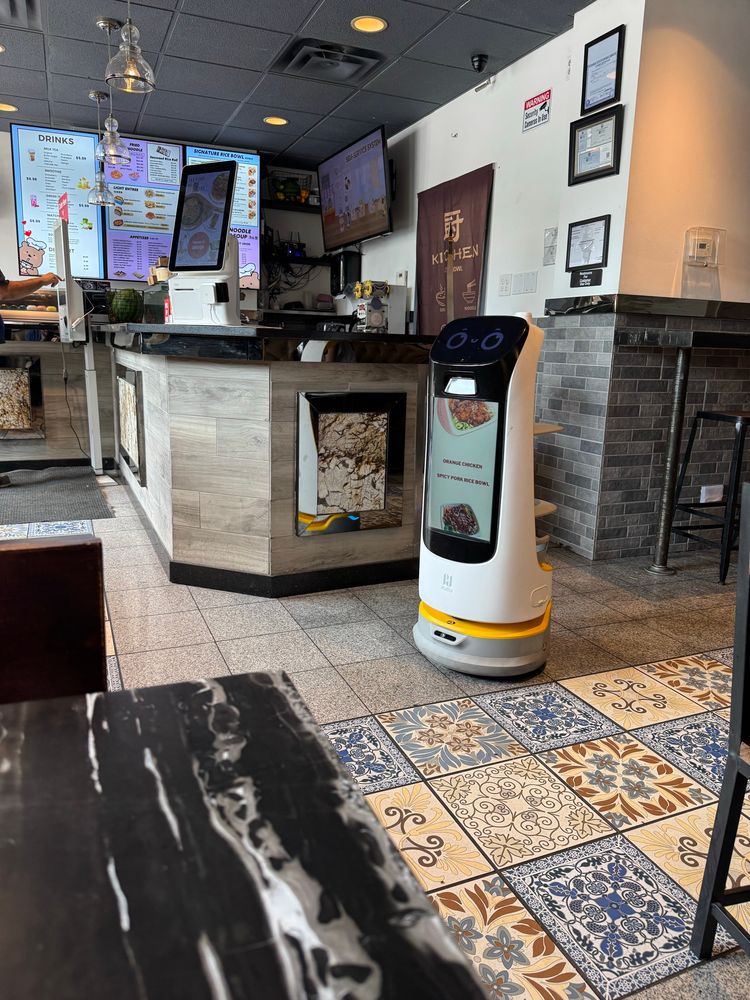 An indoor restaurant scene featuring a robotic server next to the counter. The counter displays menus and electronic devices, with a decorative tiled floor at BellyBowl Kitchen and Boba Bar in South Loop