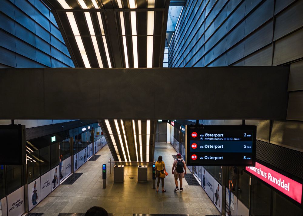 A modern subway station interior with sleek metallic walls and bright lighting. Two people are seen walking towards a digital information board displaying train schedules, including directions to Østerport. The area features glass barriers and promotional posters along the walls.