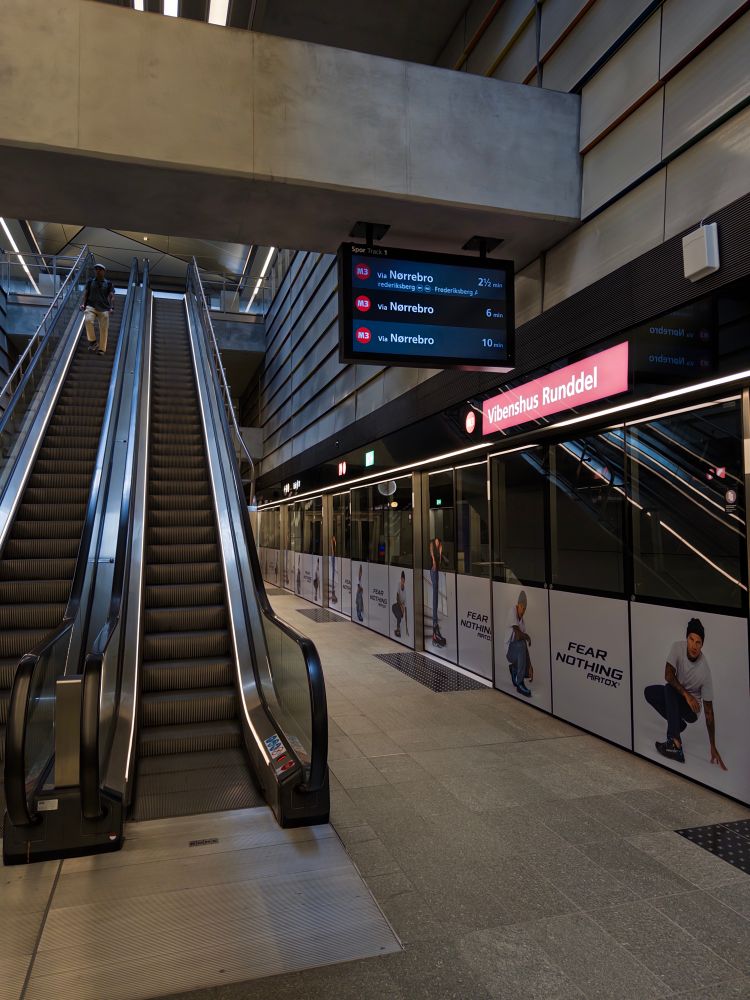 An escalator leads down to a modern subway station, featuring digital screens displaying train information. The station signs indicate routes to Nørrebro. Promotional banners line the walls, featuring athletic imagery with the slogan "FEAR NOTHING." The overall design is sleek