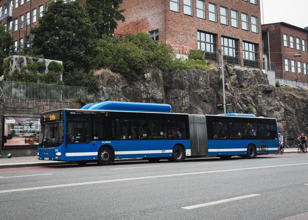 A blue articulated bus is parked on a street, with a rocky hillside and greenery in the background. Nearby, a building with a red brick facade is visible, along with an advertisement poster. Cyclists ride on the road beside the bus.