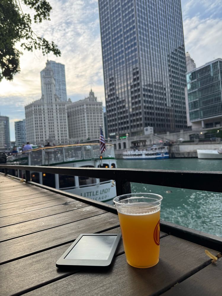 A beer and an ereader sit on a wooden table overlooking a lake. There are large tour boats in the water with tall skyscrapers in the background
