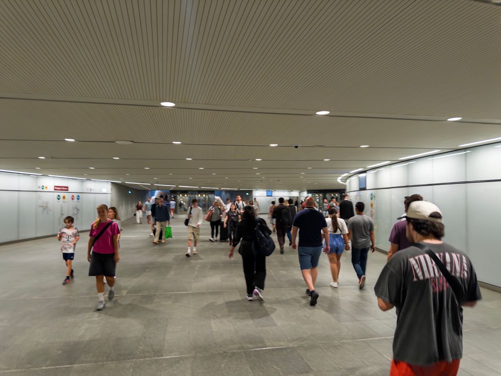 A bustling underground corridor filled with pedestrians. People of various ages are walking in both directions, with some carrying bags. The environment is well-lit, featuring a modern design with signs on the walls.
