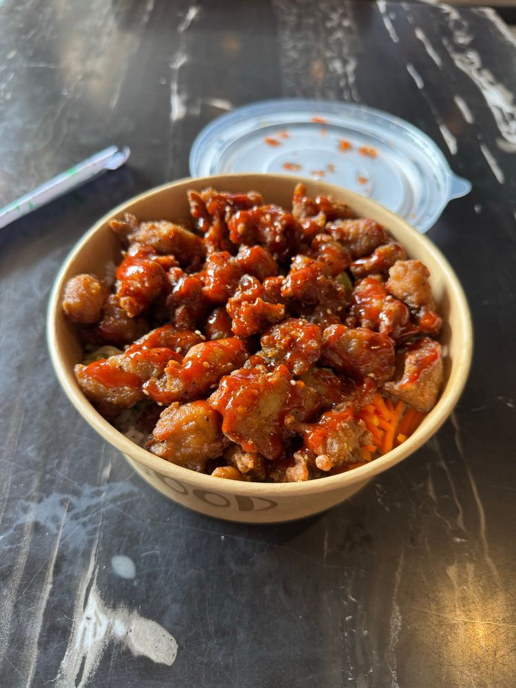 A bowl filled with fried chicken pieces coated in a red sauce, served on a bed of shredded carrots. A clear lid is partly covering the bowl, and a disposable utensil is nearby on a dark, marble surface.
