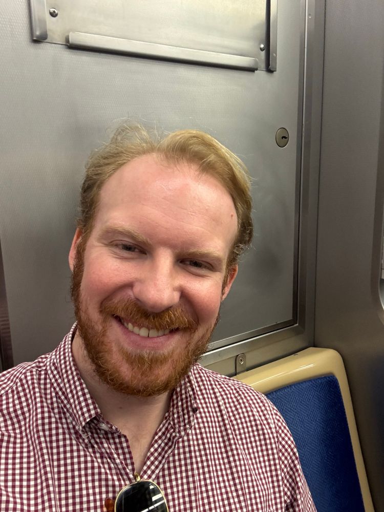 A smiling man with a reddish beard and blond hair is taking a selfie in a subway train. He is wearing a checkered shirt with sunglasses hanging off the middle. The background includes metallic walls and a blue fabric seat.