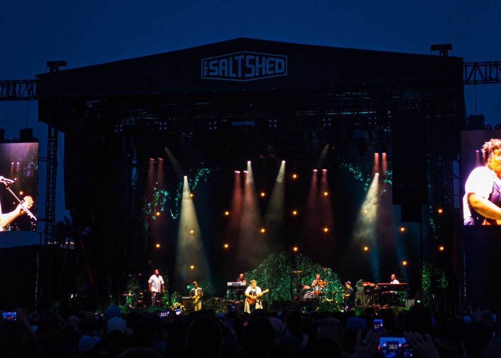 A live music performance at an outdoor venue called The Salt Shed. The stage features musicians with instruments, colorful stage lights, and a crowd of fans. Large screens display close-ups of the performers. The setting is during twilight.