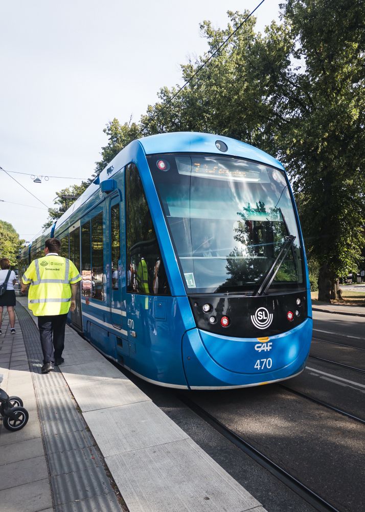A blue tram is stopped at a station with a service worker in a reflective jacket walking away. People are waiting at the platform, and trees are visible in the background.