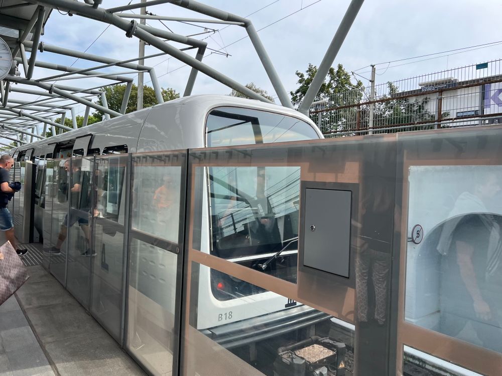 A modern train is at a station, visible through glass barriers. People are seen boarding and waiting, with a clock and overhead infrastructure in the background. The environment appears urban and well-maintained.