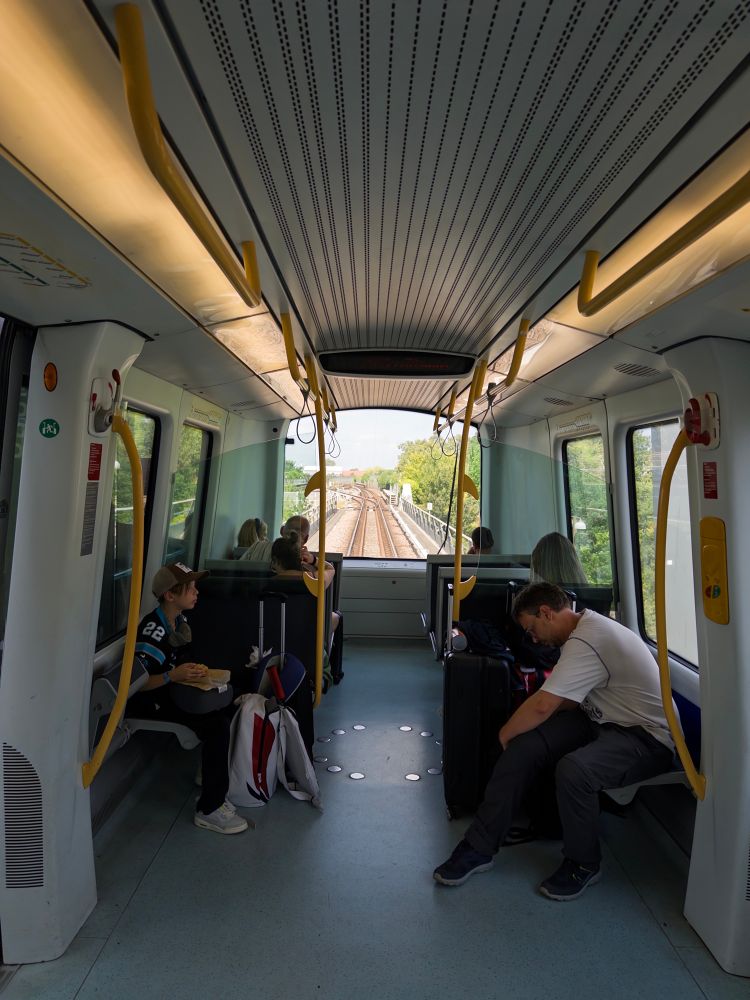 A view of the interior of a train, showing passengers seated and standing. The foreground features a child eating, while a man rests with his head down. The train windows reveal tracks and greenery outside. Yellow handrails line the ceiling, and overhead lights