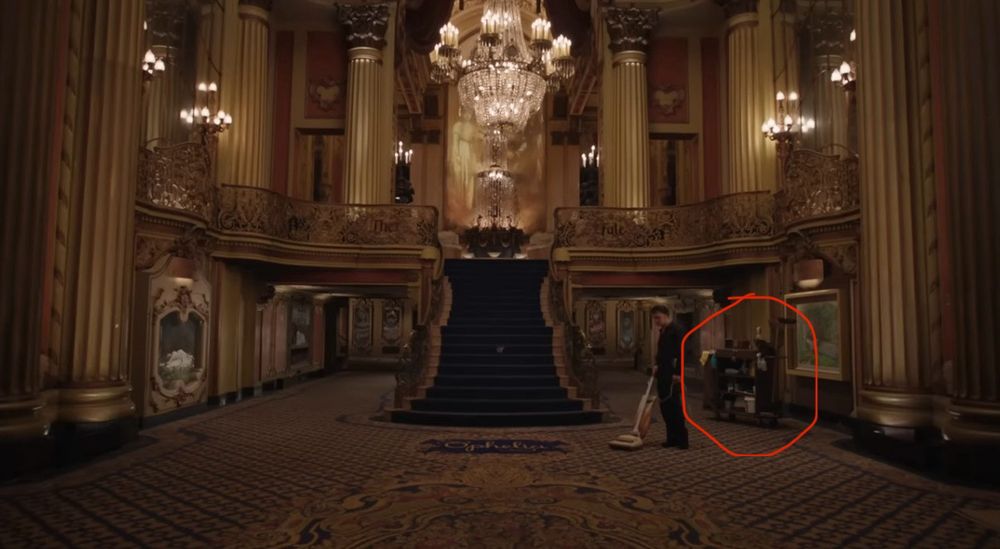 A screenshot of the first scene of the fate of Ophelia music video of Taylor swift: it depicts a grand lobby with a grand staircase, chandelier, and a guy dressed in black vacuuming the floor with a cleaning cart behind him that I circled in digital red pen