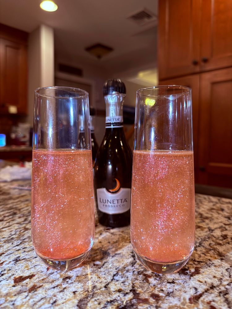 A photo of 2 stemless champagne glasses with prosecco in them but the prosecco is pink and sparkly because I put edible glitter in them and then a small bottle of prosecco is in the background