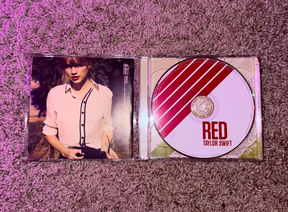 A photo of the Red (2012) CD (opened) that has a photo of Taylor in a collared shirt with the sleeves rolled up to her elbow (on the left) and on the right is the actual CD and the background is my carpet 
