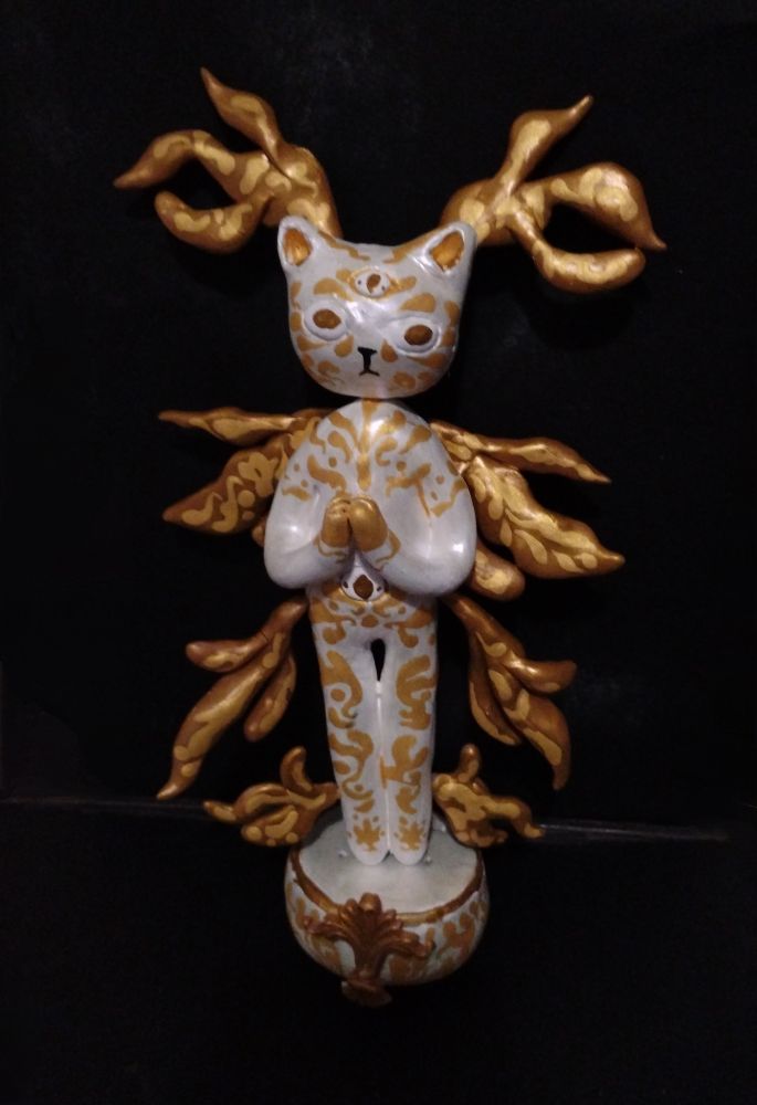 A polymer clay sculpture of an anthropomorphic angelic cat. They have white fur with gold Rococo inspired markings covering their body. With four pairs of golden twisting wings. They have four golden eyes. Three on their head and one on their stomach. 