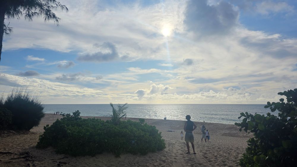 A view of the beach on top of a hill with a cloudy sky and sun shining brightly. Beachgoers on the sand walking around or enjoying the view. 