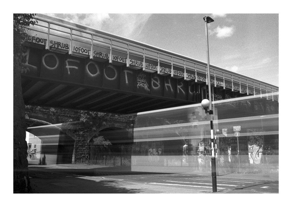 Black and white long exposure photograph of a railway bridge, the blur of a bus driving under the bridge with the blur of a train going over the bridge. 10Foot and Shrub tags cover the bridge.