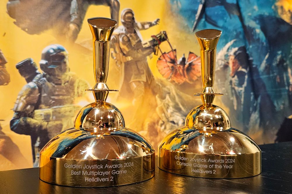 The Golden Joystick Best Multiplayer Game trophy and the Console Game of the Year trophy on a table in front of a Helldivers 2 backdrop.