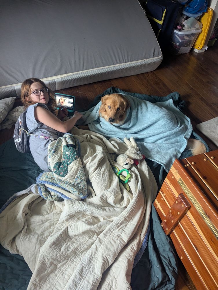 Little girl with brown hair and purple glasses, wearing a grey t-shirt and a grey feeding tube pump backpack, is laying on a grey blanket. She's covered up by a 'Sun Bonnet Sue' quilt made by her great-grandma. Next to her is a golden retriever wrapped up in a light blue baby blanket. They're looking at the camera, slightly bothered that their tablet time has been interrupted for a quick picture. 
