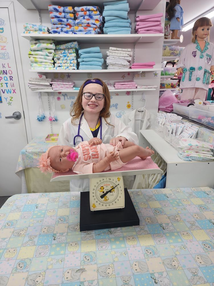 Little girl dressed in a white doctor's coat, laying a baby doll on top of a baby scale, surrounded by baby blankets and diapers. 