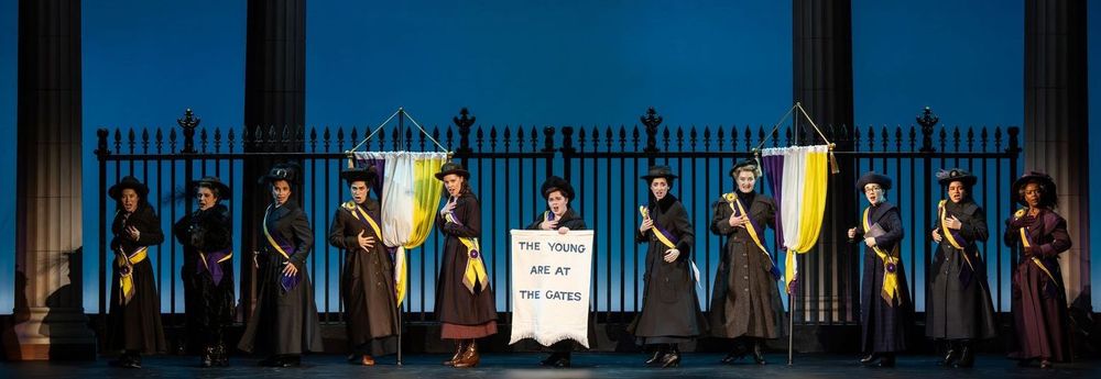 The all female cast in front of a long steel fence and a handheld sign, "The young are at the gates" in the first national touring company of the musical, “Suffs”.  Photo courtesy of Joan Marcus