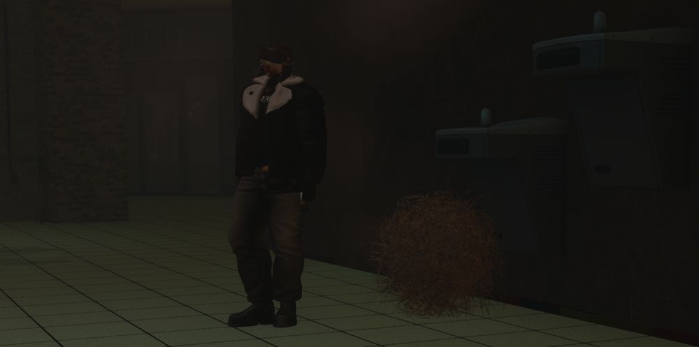 A tall, heavily dressed person standing inside what looks to be a dimly lit mall, stood beside two strangely large drinking fountains. In between the person and the fountains is a tumbleweed. It somehow looks very fitting to the atmosphere of the setting, despite being inside a mall.