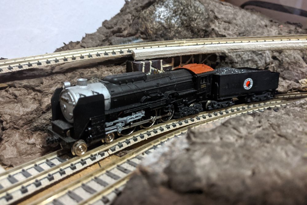 A model train locomotive in a bare model railroad landscape, with terrain forms made from brown-coloured papier-mâché but nothing covering them. The locomotive is a Japanese class C62, which should be all-black, but someone has painted the smokebox silver, the cabin roof brown, and added a logo to the side of the tender.