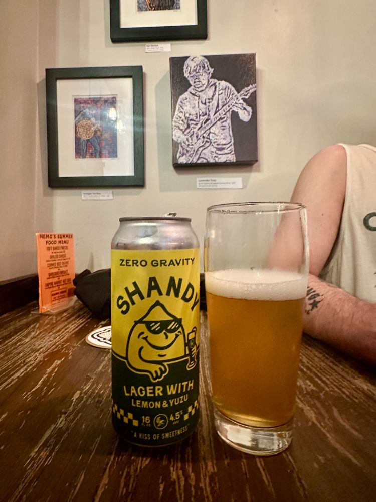 Shandy beer can next to my glass of beer and oomfs arm to the right. On the wall is artwork including Trey on a guitar