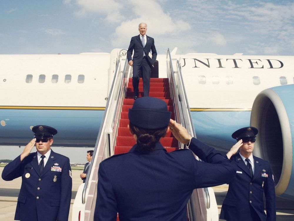 Beautifully shot and staged photo of President Biden exiting a plane. In the foreground is the back of a female air force officer (forgive me for knowing nothing of titles or ranks, she is wearing a navy blue uniform and cap) saluting. To either side of the stairs leading out of the plane there are two male air force officers facing us, also saluting. 