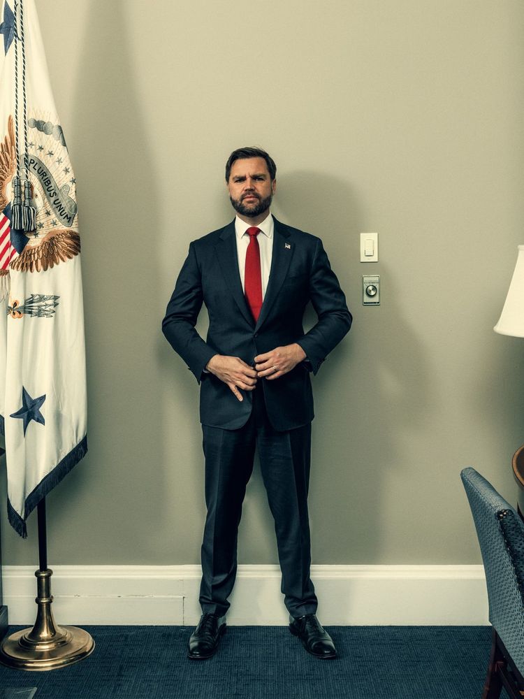 Full body portrait photograph of JD Vance, a smarmy-looking thirtyish man in a dark suit with a red tie and white shirt. He is posed against a blank gray wall. To his right is a flag on a flagstand, only 2/3s of it in shot. Next to his left upper arm on the wall is a light switch and a thermostat. On the left side of the shot, we can see the very back of a blue chair in side profile, a bit of a lampshade and a tiny sliver of a side table.