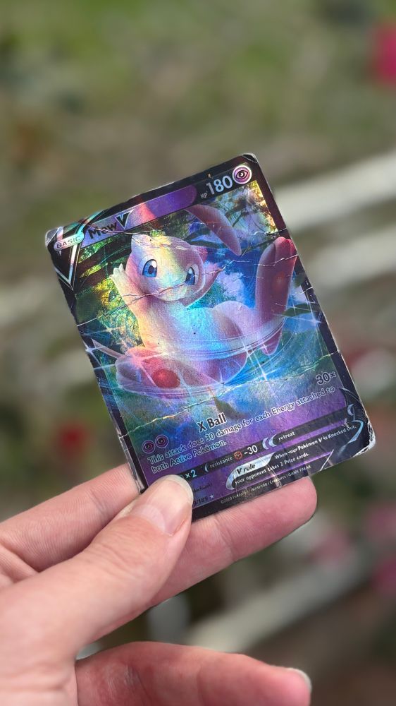 a slightly worn shiny Pokémon card of mew, glimmering softly in the morning light