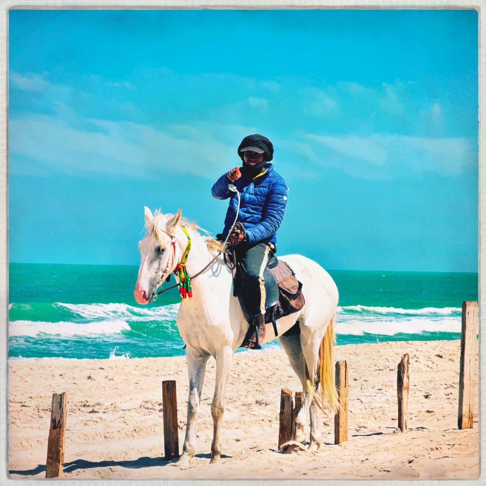 Mauritanian man barefoot on a white horse on the beach.  Blue Sky and sea in the background on a windy day