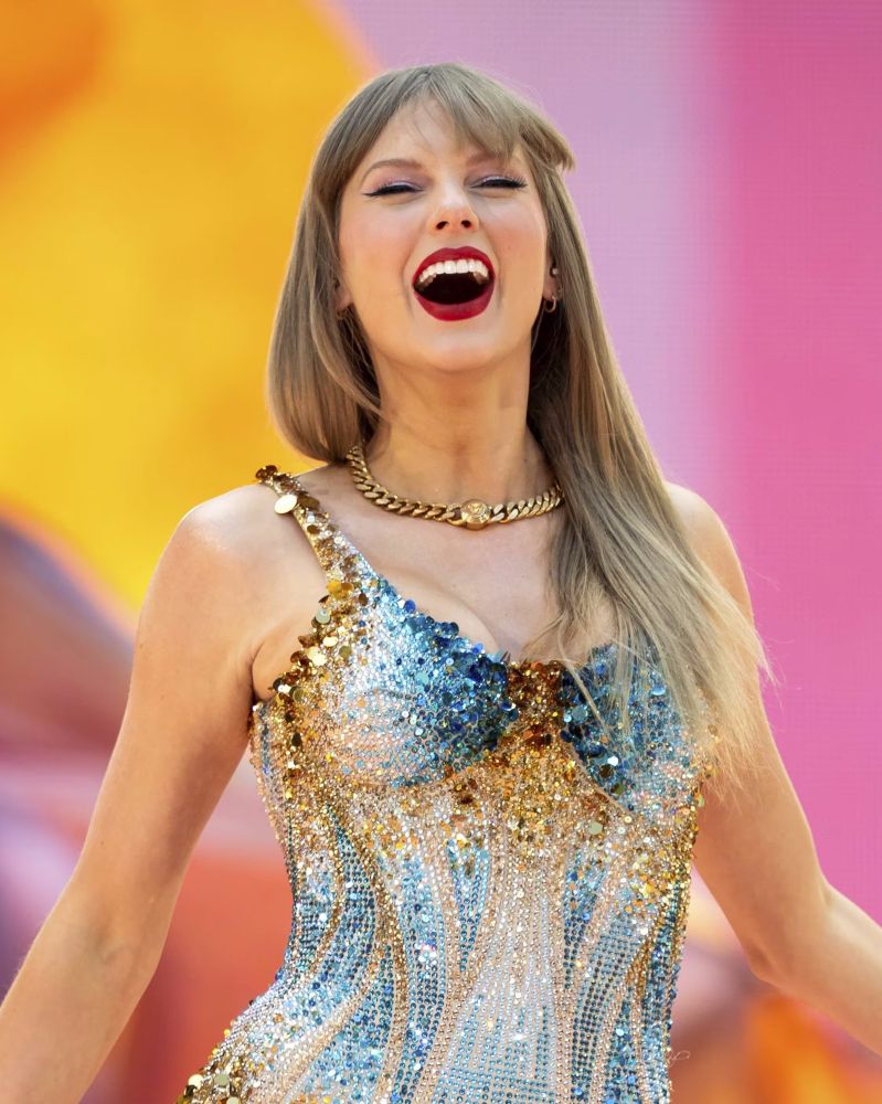 Taylor Swift at The Eras Tour during Lover era. She is wearing the gold and blue bodysuit. She shows the biggest smile that lights up her face in a beautiful way. She looks so happy.