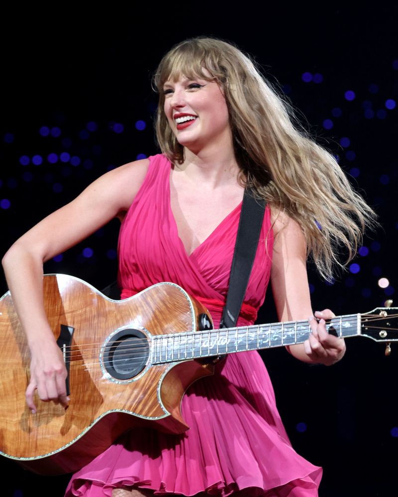 Taylor Swift at The Eras Tour during the acoustic set. She’s wearing the pink dress and has her guitar in her hands. Her face is brightened by the cutest smile and looks super happy.