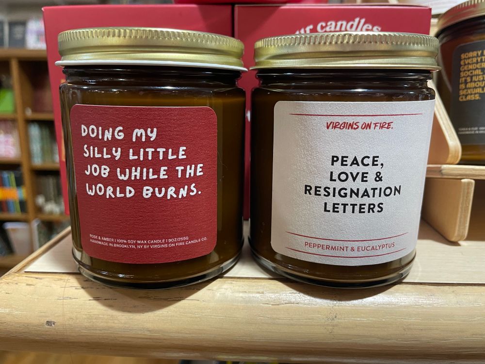 Two mason jar candles from Virgins on Fire. 

The one on the left has a red label, rose and amber scent, and is named: “DOING MY SILLY LITTLE JOB WHILE THE WORLD BURNS”

The one on the right has a white label, peppermint & eucalyptus scent, and is named: “PEACE, LOVE & RESIGNATION LETTERS”