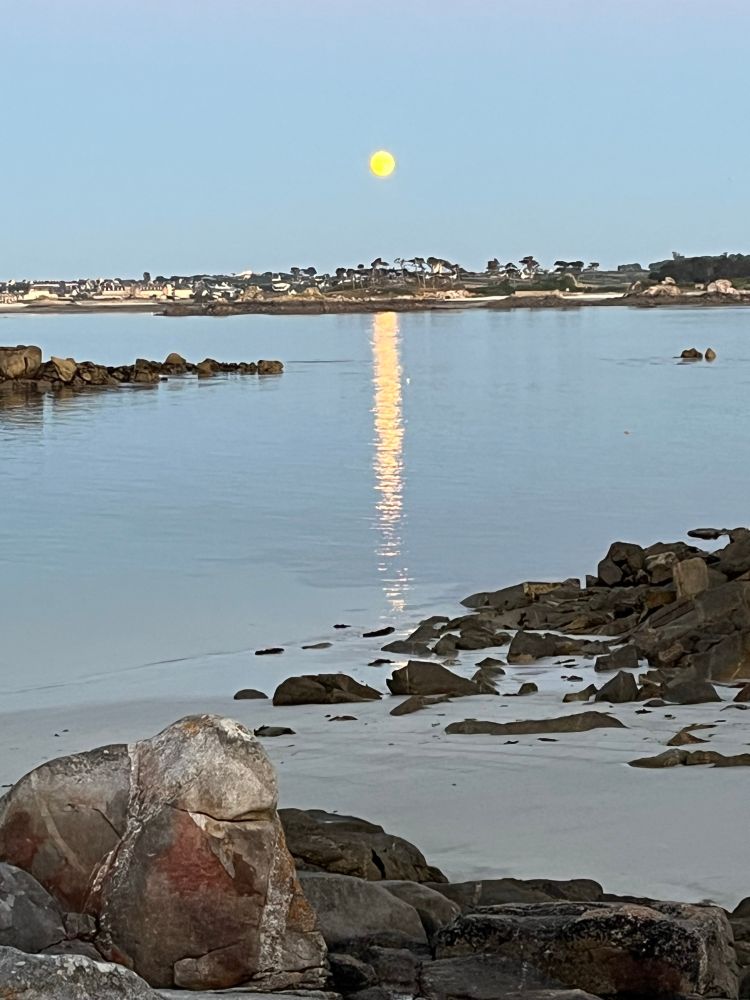 Lever de pleine lune sur la mer, au premier plan quelques rochers et un peu de sable découverts par la marée basse, puis un beau reflet de la lune sur l’eau calme, au loin la côte de Roscoff - Finistère, ciel sans nuages et pleine lune