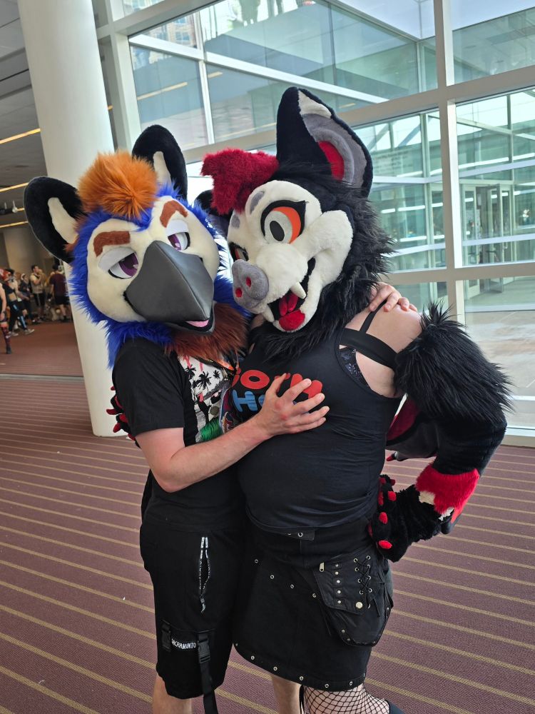 A bat fursuiter standing next to a bird fursuiter while the bird squeezes the bat's chest rather suggestively!