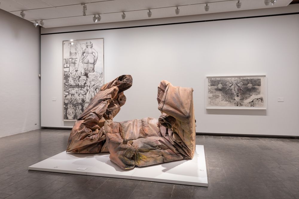 A gallery view that features drawings hung on the walls and a central sculpture that consists of two folded and crushed leather sofas.
