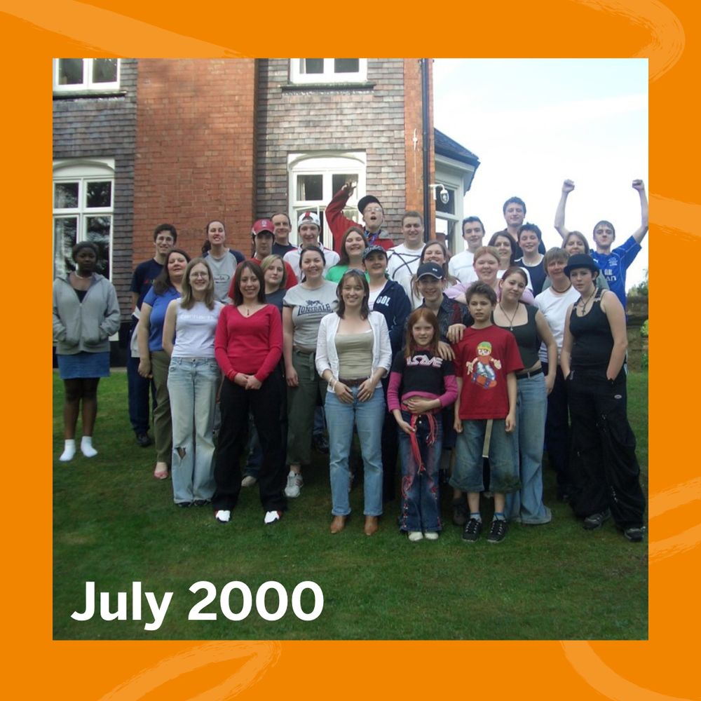 A large group of people posing outdoors on grass in front of a brick building with white-framed windows in July 2000.