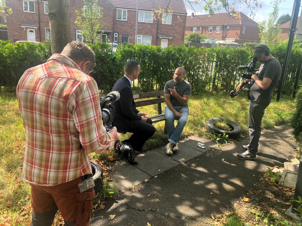 Two men sit on a bench in a residential area while two cameramen film them outdoors on a sunny day.