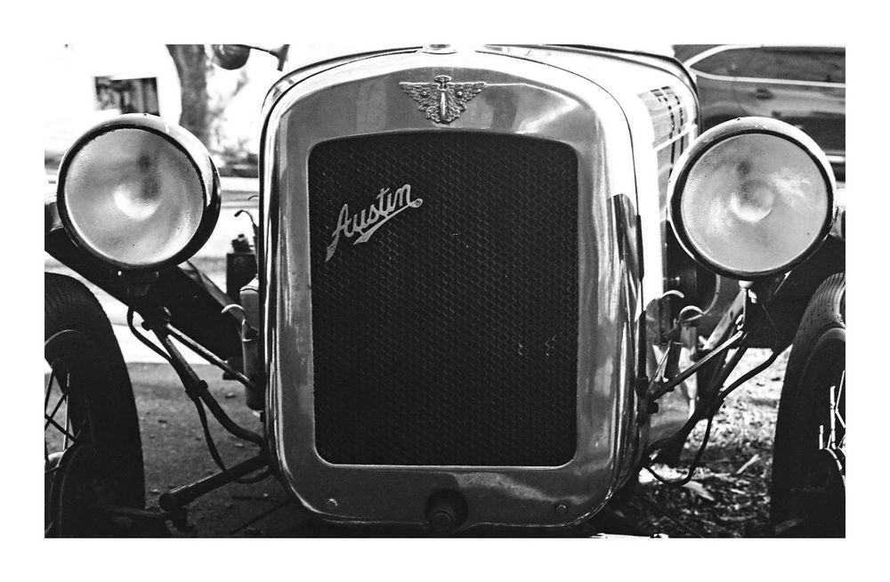 A close up of the front of an Austin 7 vintage car.  In black and white. You can see the grill, headlights and some of the front wheels. 