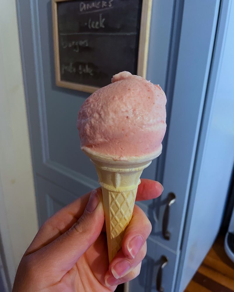 A hand holding an ice cream cone in front of a blue fridge with a scoop of strawberry ice cream in it