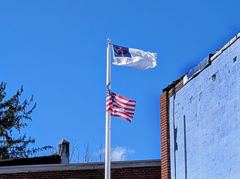 A Christian flag (white with a blue rectangle in the upper left which has a red cross on it) flies above the American flag on a sunny day.