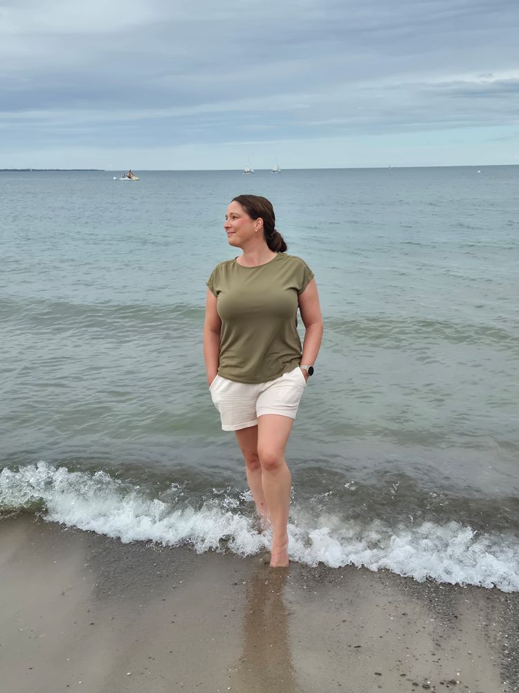 Nina steht in hellen Shorts und grünem T-Shirt am Strand, mit den Füßen im Wasser, Hände in den Taschen. Blick nach links lächelnd. Dahinter nichts als flaches grau-grünes Meer und ein grau-grün-blauer Himmel.