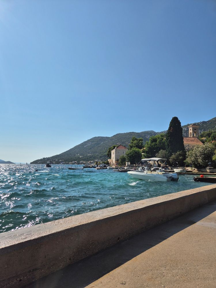 Bucht vor Viganji. Wasser, Böötchen, Kirche, Häuser, Hügel, viel blauer Himmel.